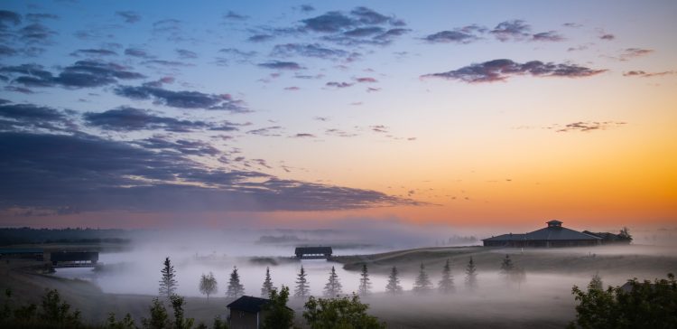 A sky with clouds and the orange glow of a sunrise over a foggy lake and events centre.