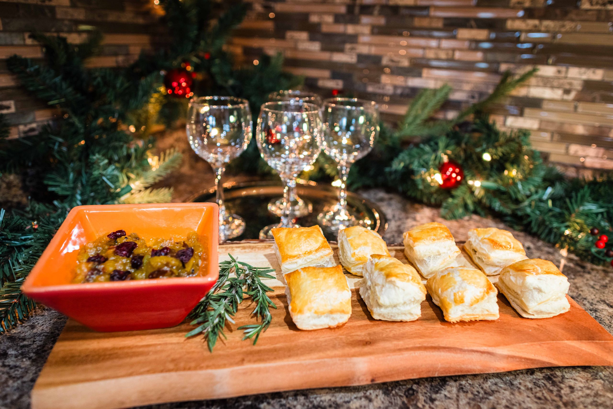 Mini puff pastries elegantly presented on a wooden platter, next to a side of chutney in a red dish and a sprig of rosemary. In the background, a twinkling Christmas garland adds festive decor to the counter