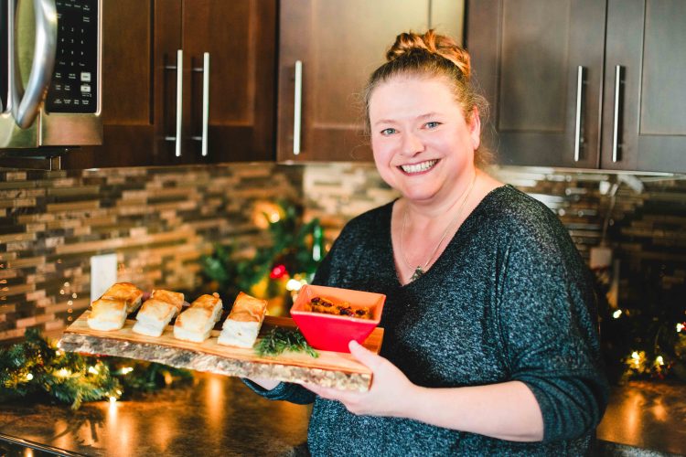 Kathy Neiman stands in her festive kitchen holding a platter of delicious tourtière puffs. A red dish of chutney complements the appetizers, and Christmas decorations twinkle in the background, creating a warm and inviting atmosphere.