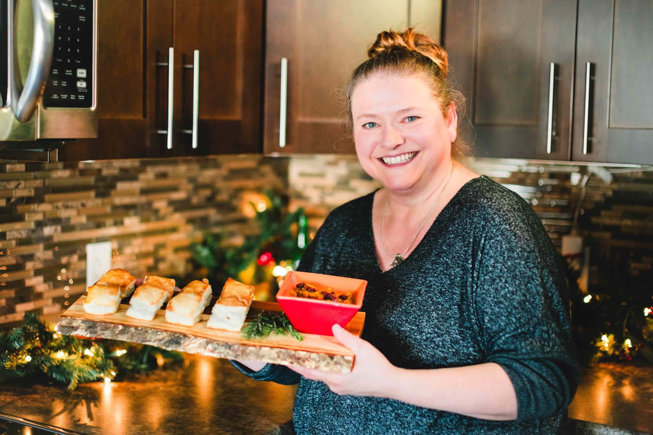 Kathy Neiman stands in her festive kitchen holding a platter of delicious tourtière puffs. A red dish of chutney complements the appetizers, and Christmas decorations twinkle in the background, creating a warm and inviting atmosphere.