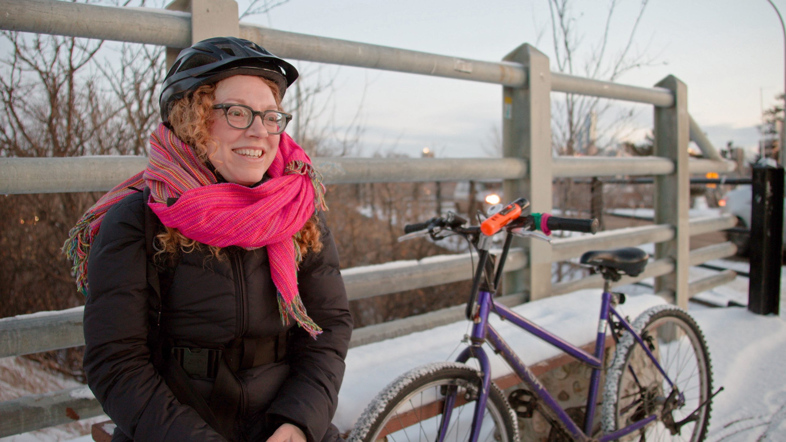 A woman wearing glasses, a pink scarf, a winter parka and a bike helmet sits on a snow-covered bench next to her purple bike.