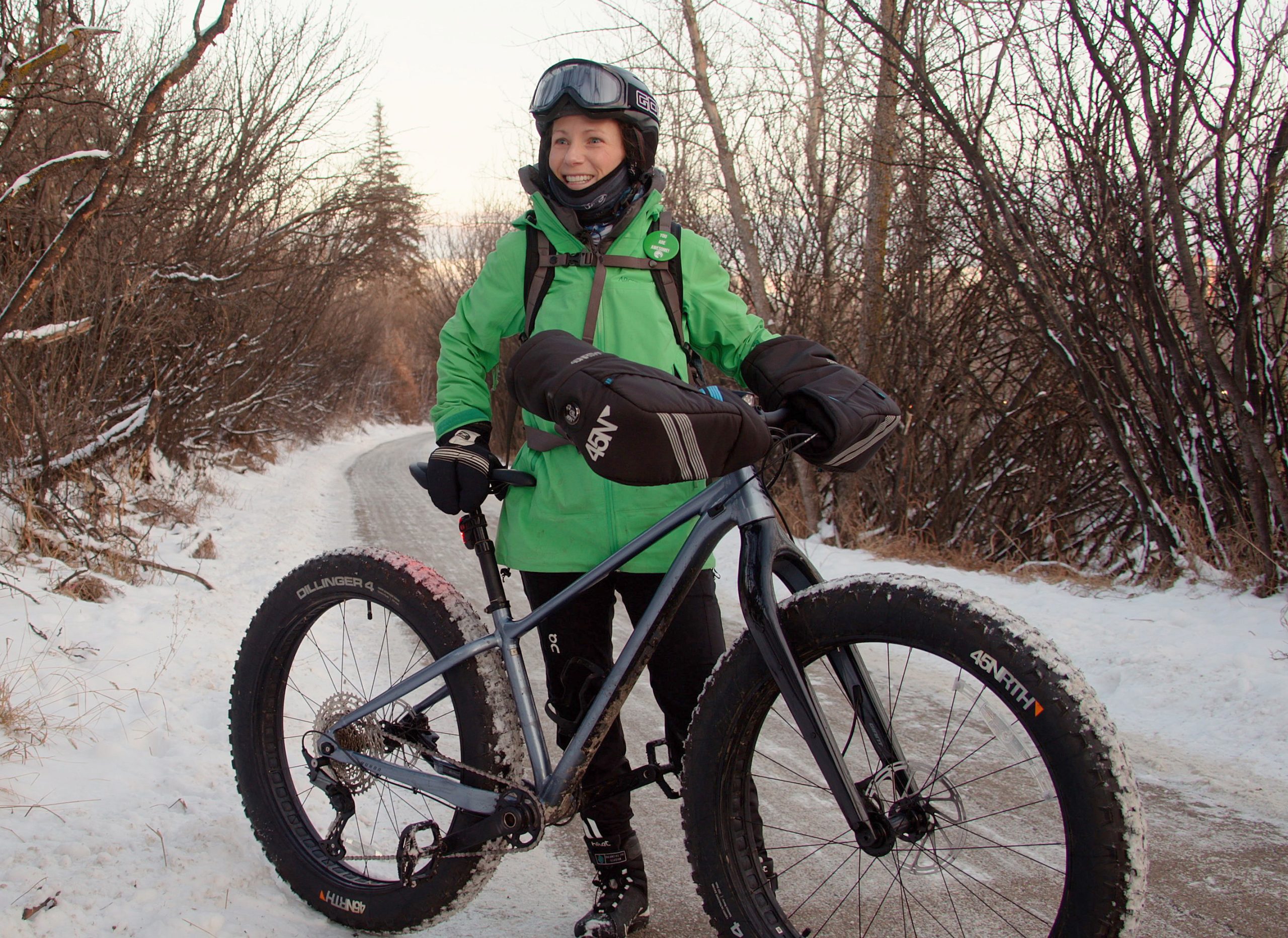 A woman wearing a green parka, bike helmet and goggles stands behind her fat bike on a trail dusted with snow.