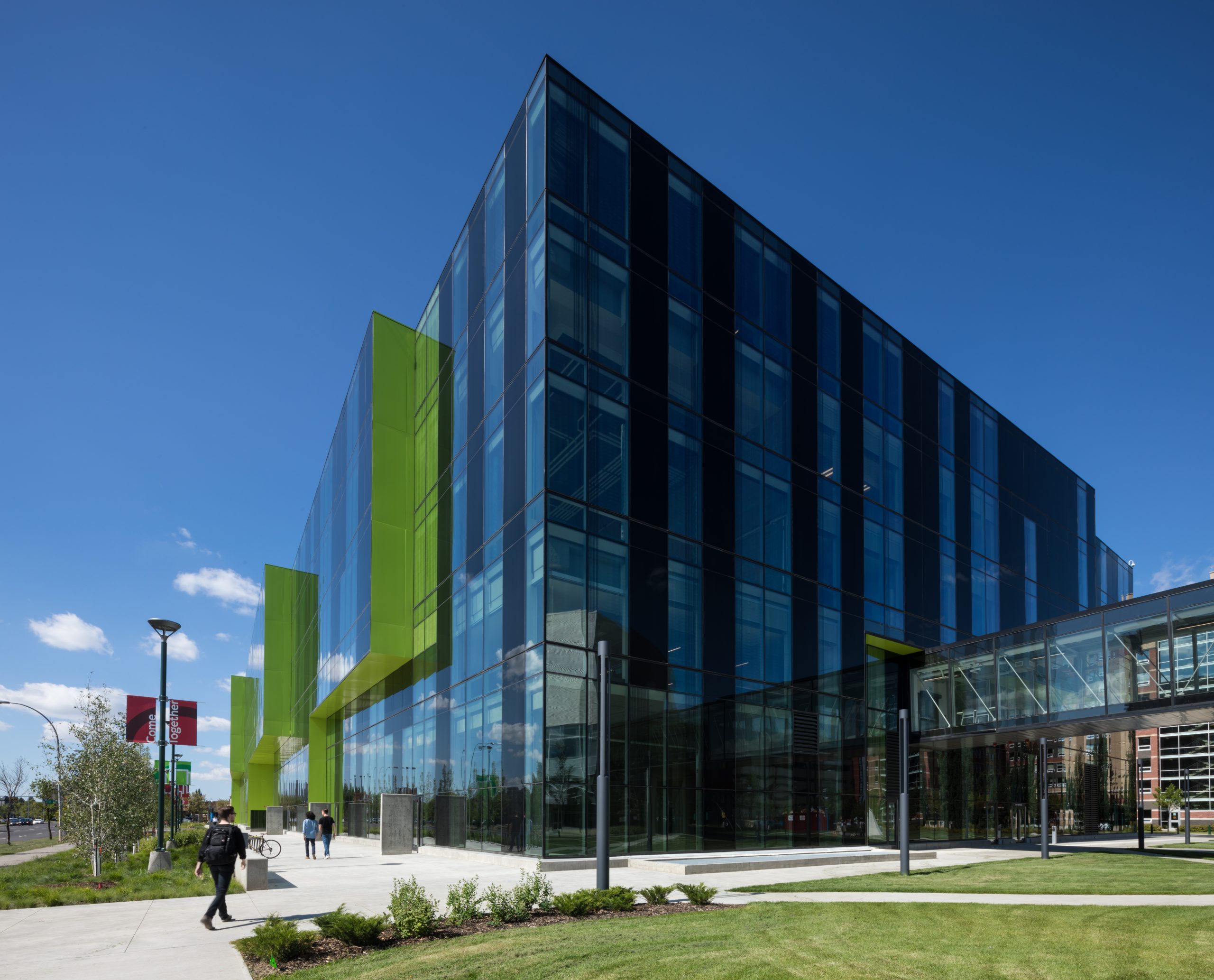 Alt-text: Three students walk along the sidewalk in front of a glass-covered building on the MacEwan University campus.