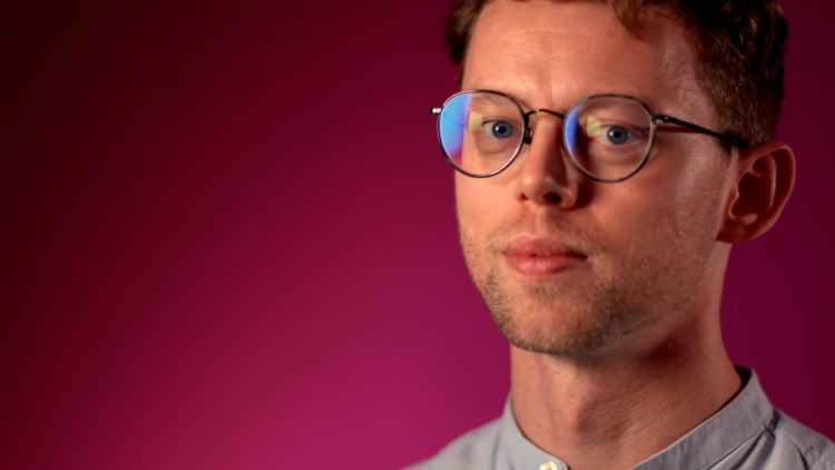 A man with glasses looking at the camera with a deep red background.