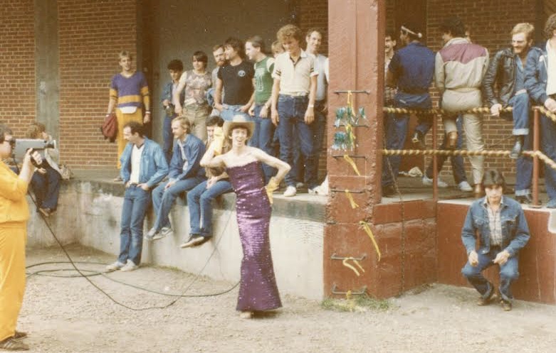 A man in a purple dress and cowboy hat poses in front of a crowd of 20 men sitting or standing on the loading dock of a building.