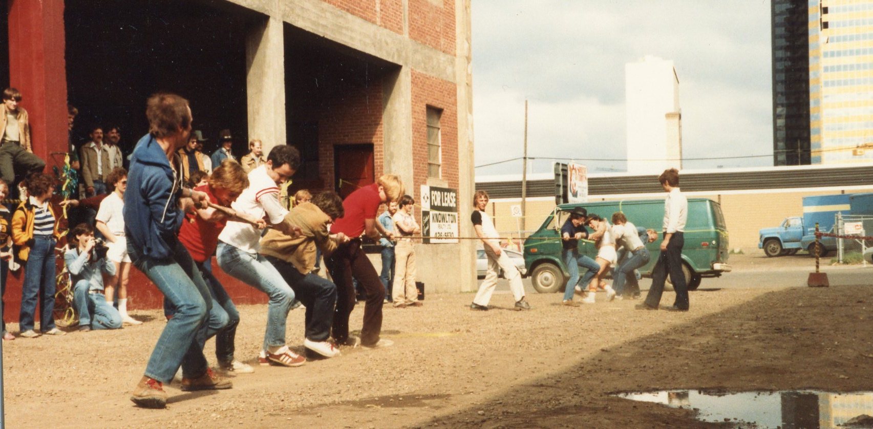 Two groups of men take part in a tug of war in a gravel alley next to a brick building.