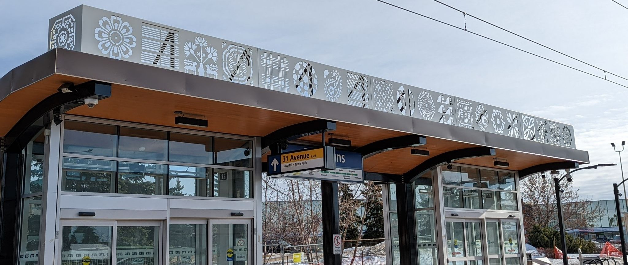 An aluminum panel, featuring multi-shaped cut-outs, runs the length of the roof of an LRT shelter like the cloth border of a blouse or a dress.