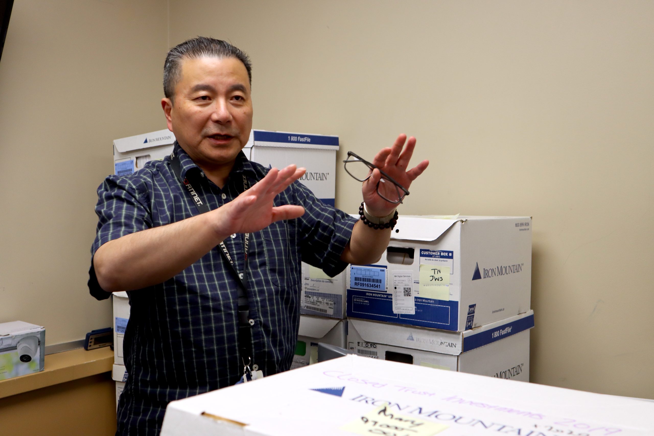 A man stands in an office with stacked boxes behind him giving instructions to someone out of frame.