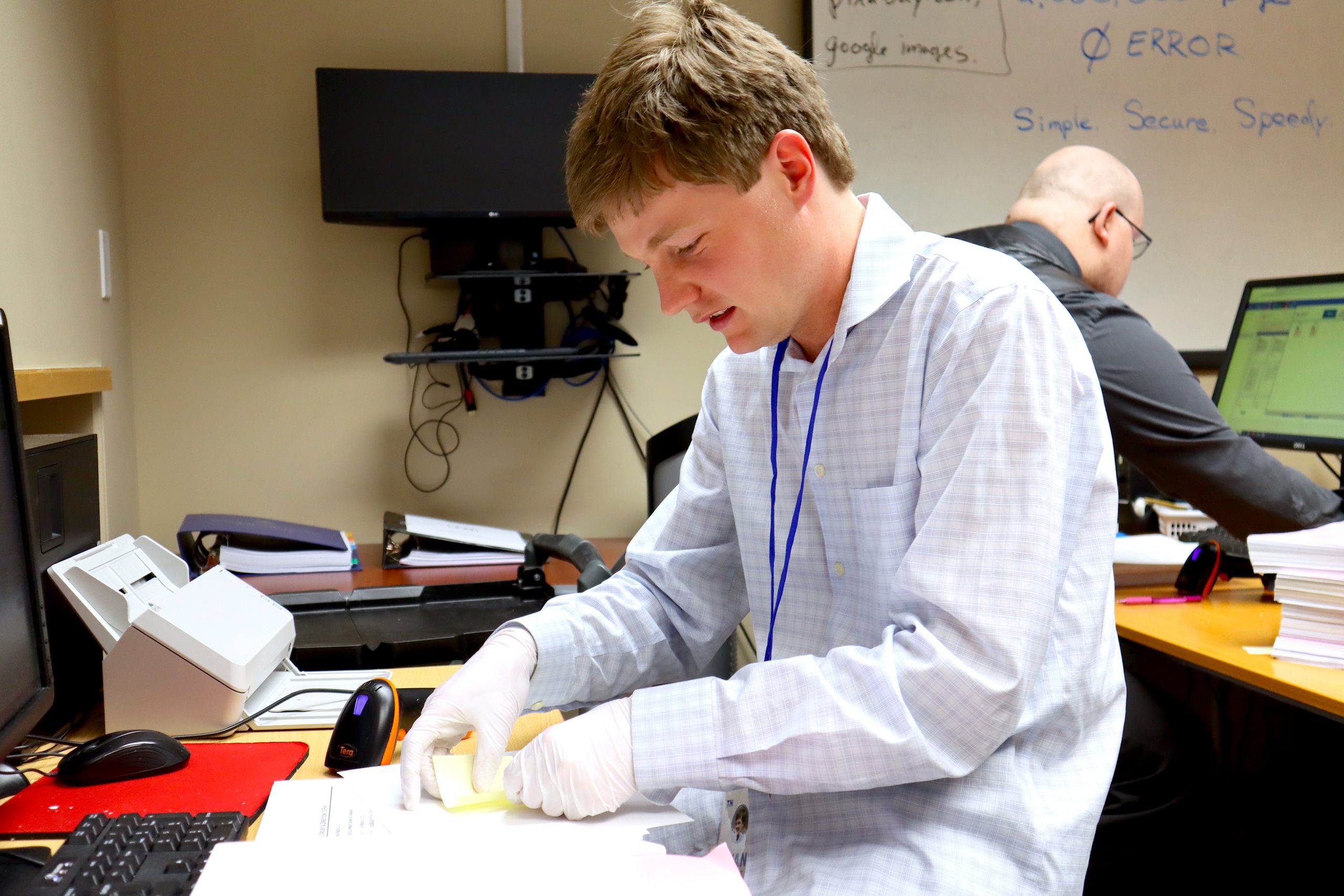 A young man handles a sticky note on a document as another man looks at a computer screen in the background.