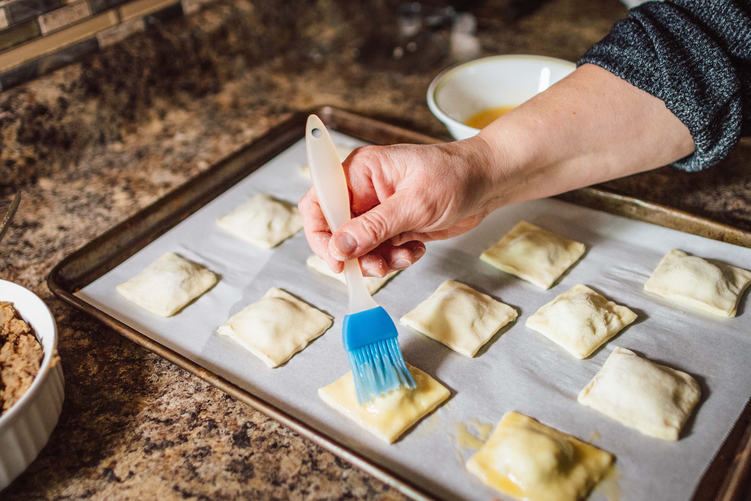 Close-up of a hand delicately brushing egg wash on top of pastry squares in the final step of the cooking preparation process.