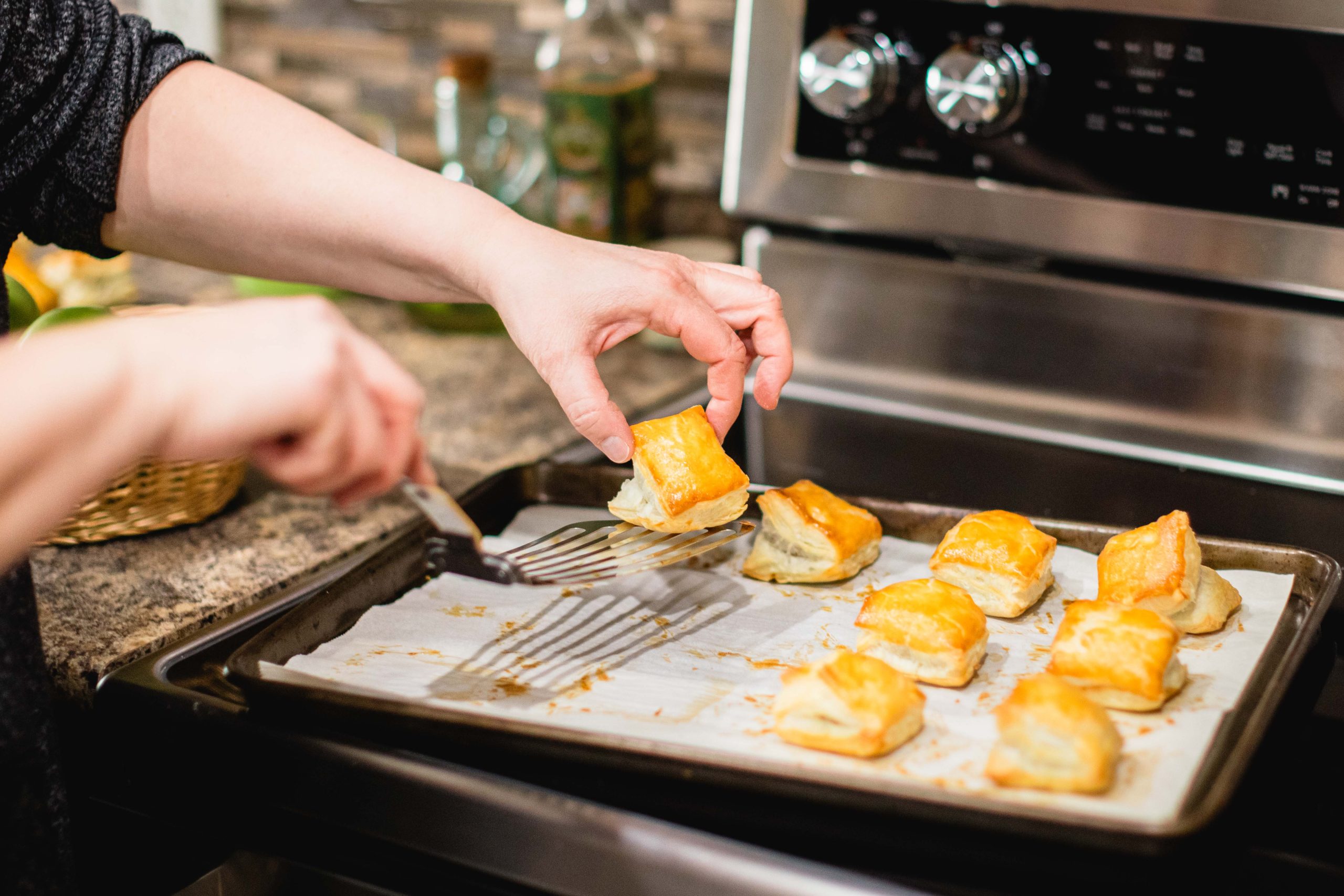 Close-up of hands using a metal spatula to carefully lift golden puff pastries from a baking tray lined with parchment paper.