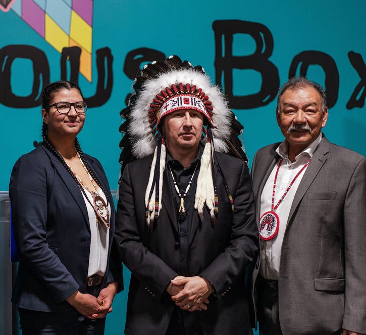 A woman stands between two men in headdresses with a third man on the right.