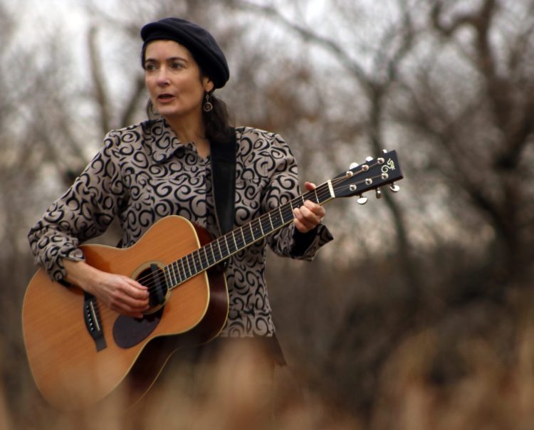 A woman, wearing a beret, plays an acoustic guitar outdoors, surrounded by trees. 