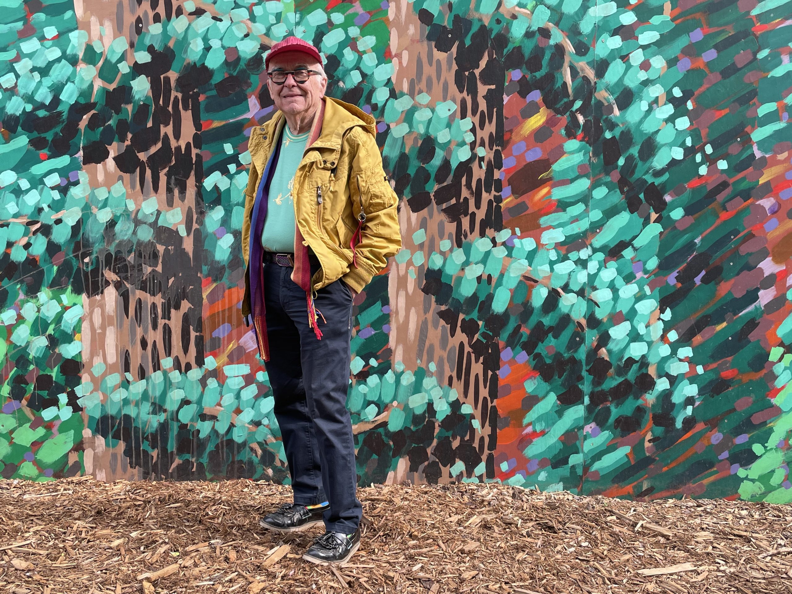 Michael Phair, dressed in a yellow jacket and red sequined ball cap, stands on some wood chips in front of a mural of trees.