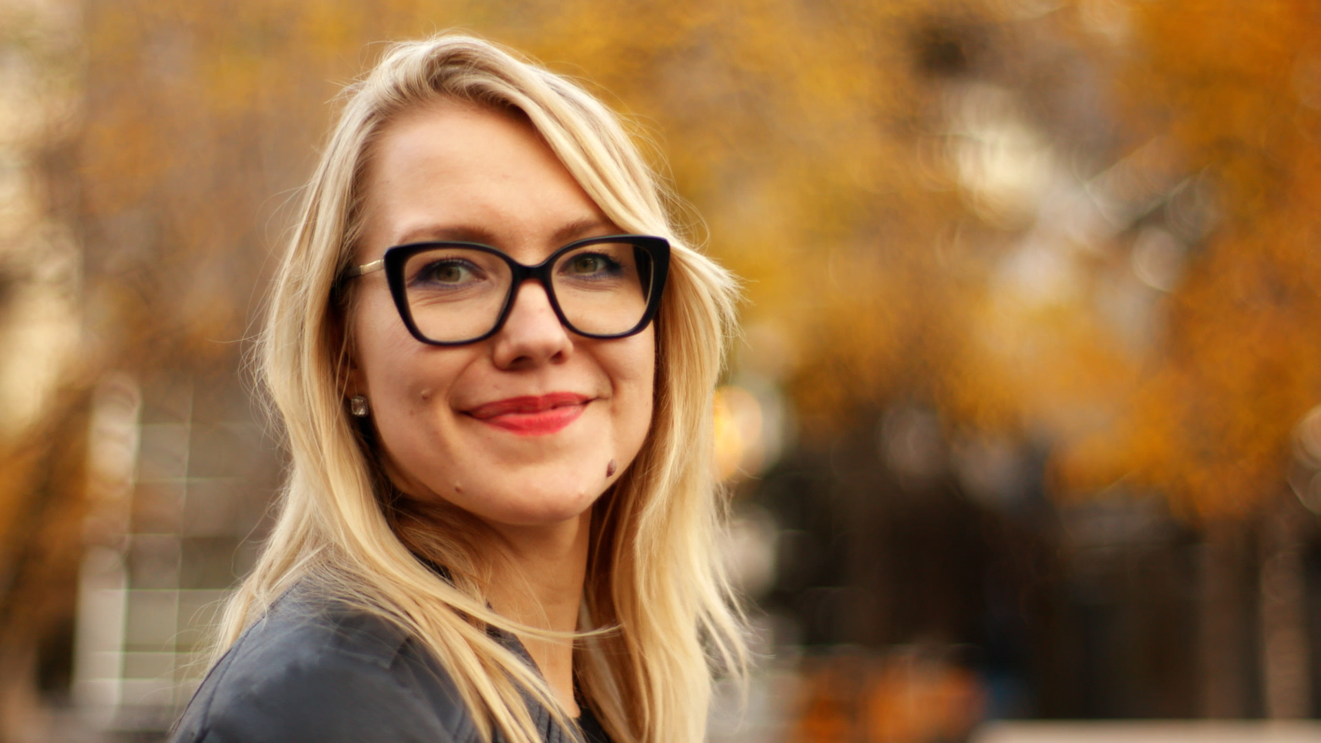 A blond woman with glasses smiles at the camera. There are yellow and orange trees in the background.