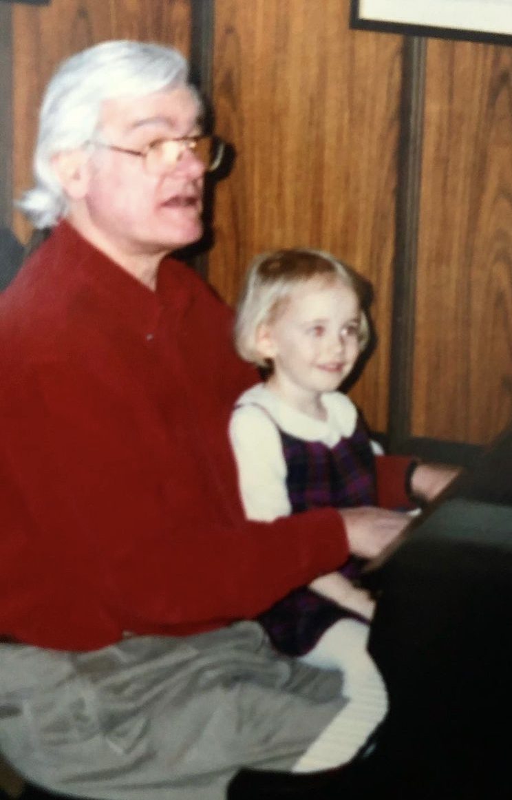 A man with white hair and glasses plays a piano with his young granddaughter on his lap.