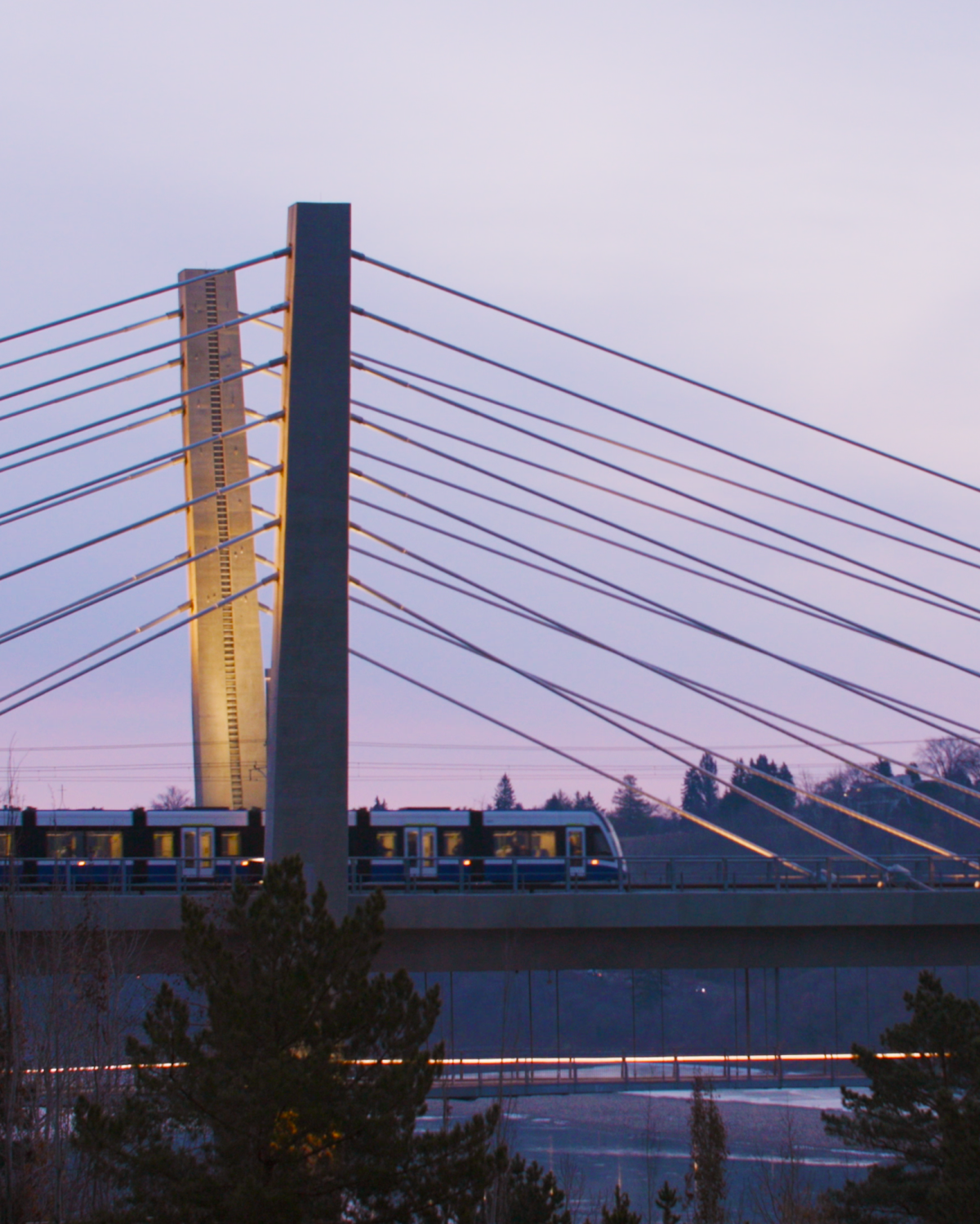 The Valley Line Southeast LRT train car, travelling across a bridge at sunrise, the cityscape in the background with warm morning light painting the sky in shades of pink and purple