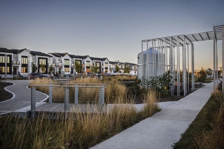 A park with green grass and walking paths occupies the foreground, featuring metal structures on the right side. In the background on the left, there are modern row houses with sleek, contemporary designs.