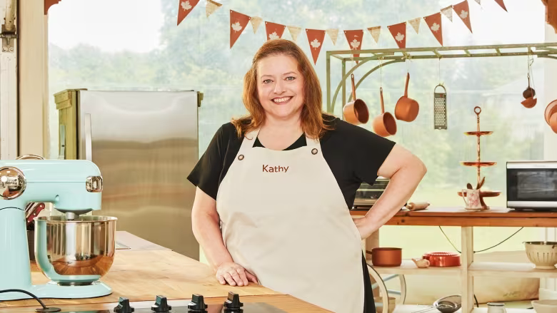 Kathy Neiman on the set of the Great Canadian Baking Show, wearing a white apron and black T-shirt. She stands at a kitchen station inside the baking tent, accompanied by a teal mixer on her side. Copper pots and pans are visible in the background. Credit: Photo courtesy of CBC.