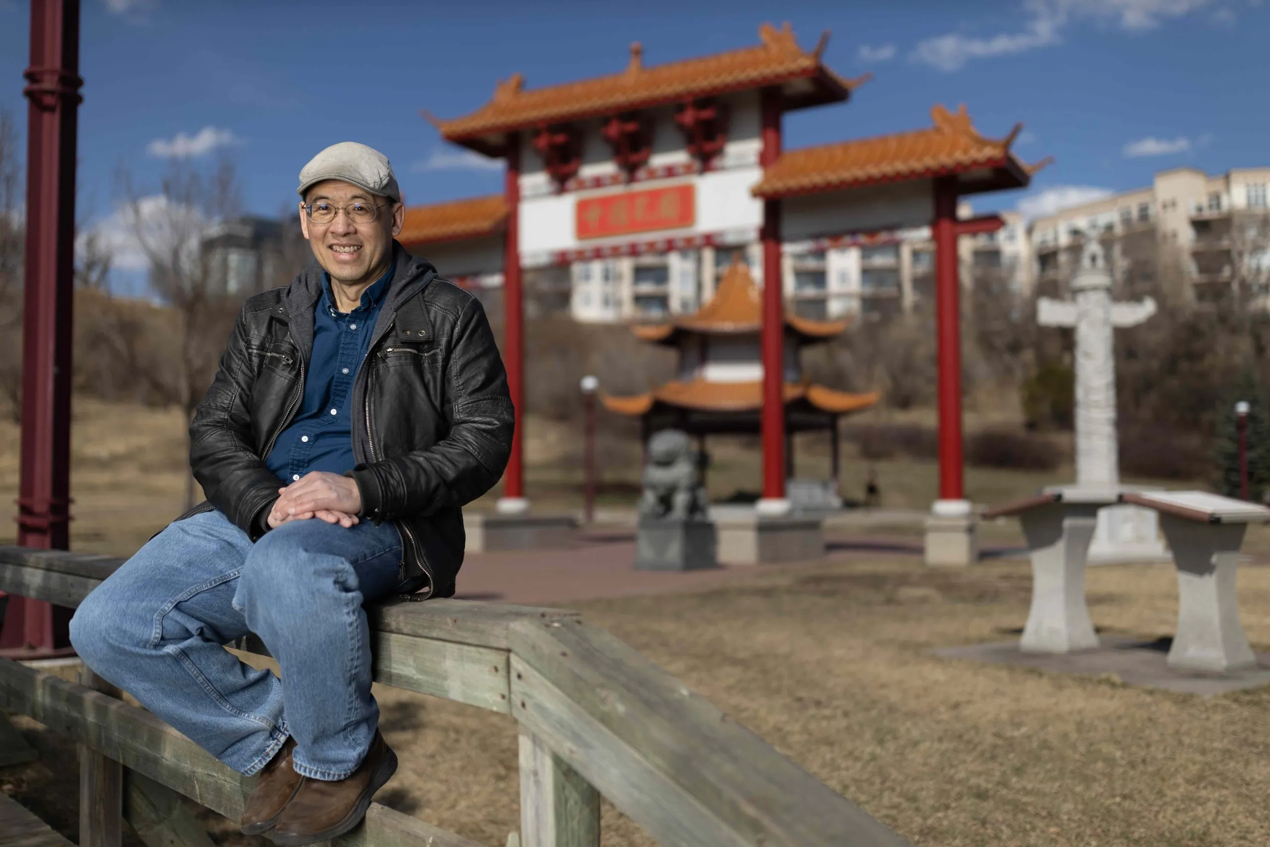 A man in a leather jacket, jeans and a cap sits on a railing in front of a Chinese gate, lion statue and pagoda.