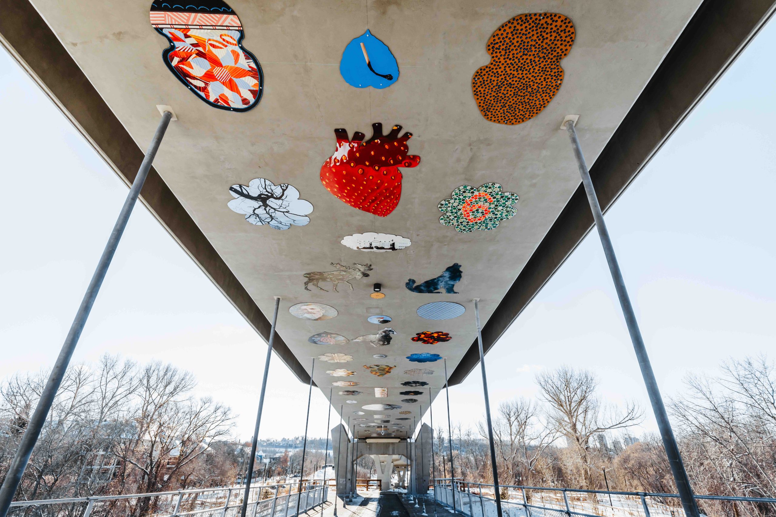 Panels of multi-coloured and multi-shaped art run the length of the ceiling of a concrete pedestrian/LRT bridge.