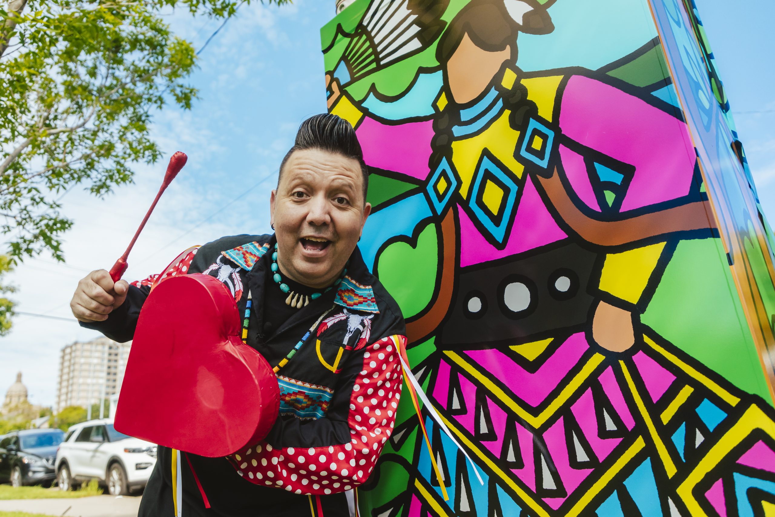 An Indigenous man holding a red stick and heart-shaped drum stands next to a traffic control box wrapped with his artwork of a jingle dress dancer holding a fan of feathers.