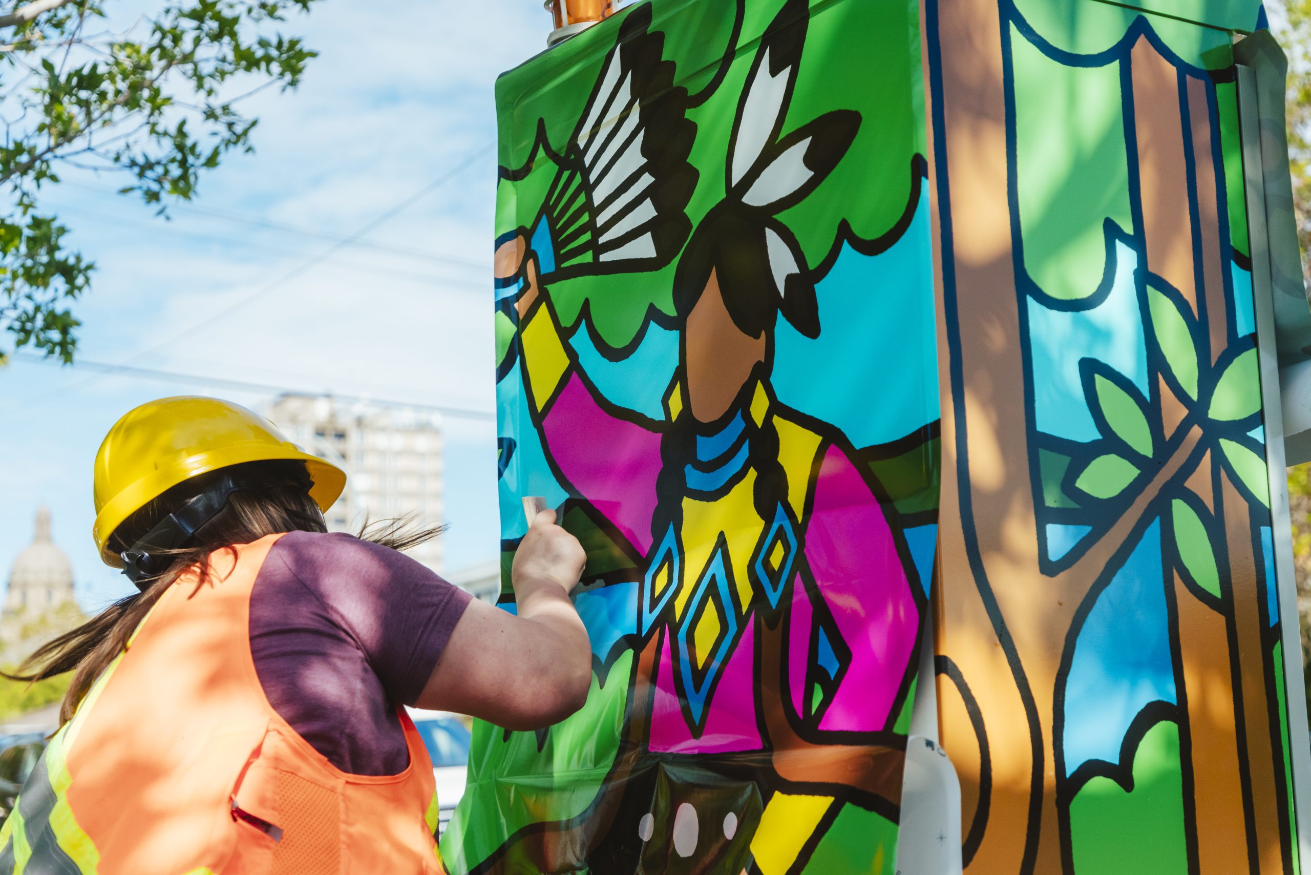 A woman in an orange vest uses a small plastic card to flatten any air bubbles as she wraps Cardinal’s art on a control box.