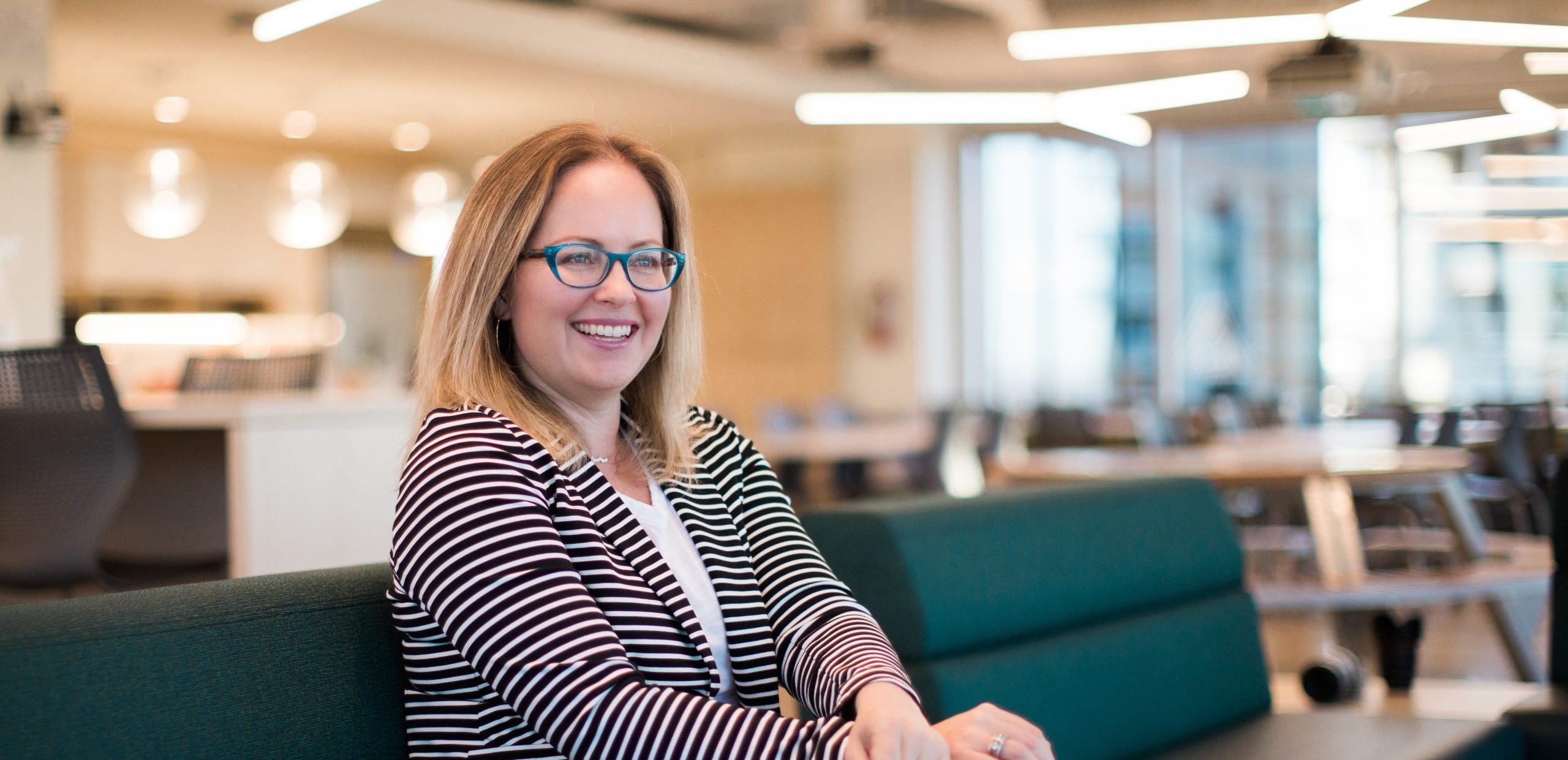 Nicole Janssen, co-founder of AltaML, sits on a green sofa in a modern office space, smiling and looking at the camera. She is wearing a striped blazer, white top, and blue jeans.