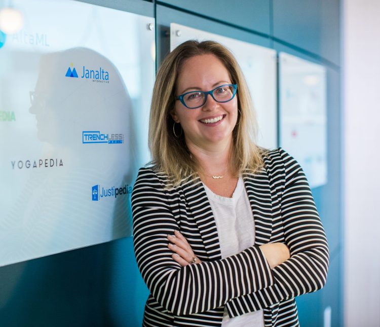 Nicole Janssen, co-founder of AltaML, stands smiling with her arms crossed in front of a glass wall displaying various company logos including AltaML, Janalta, and Yogapedia. She is wearing a striped blazer and blue jeans.