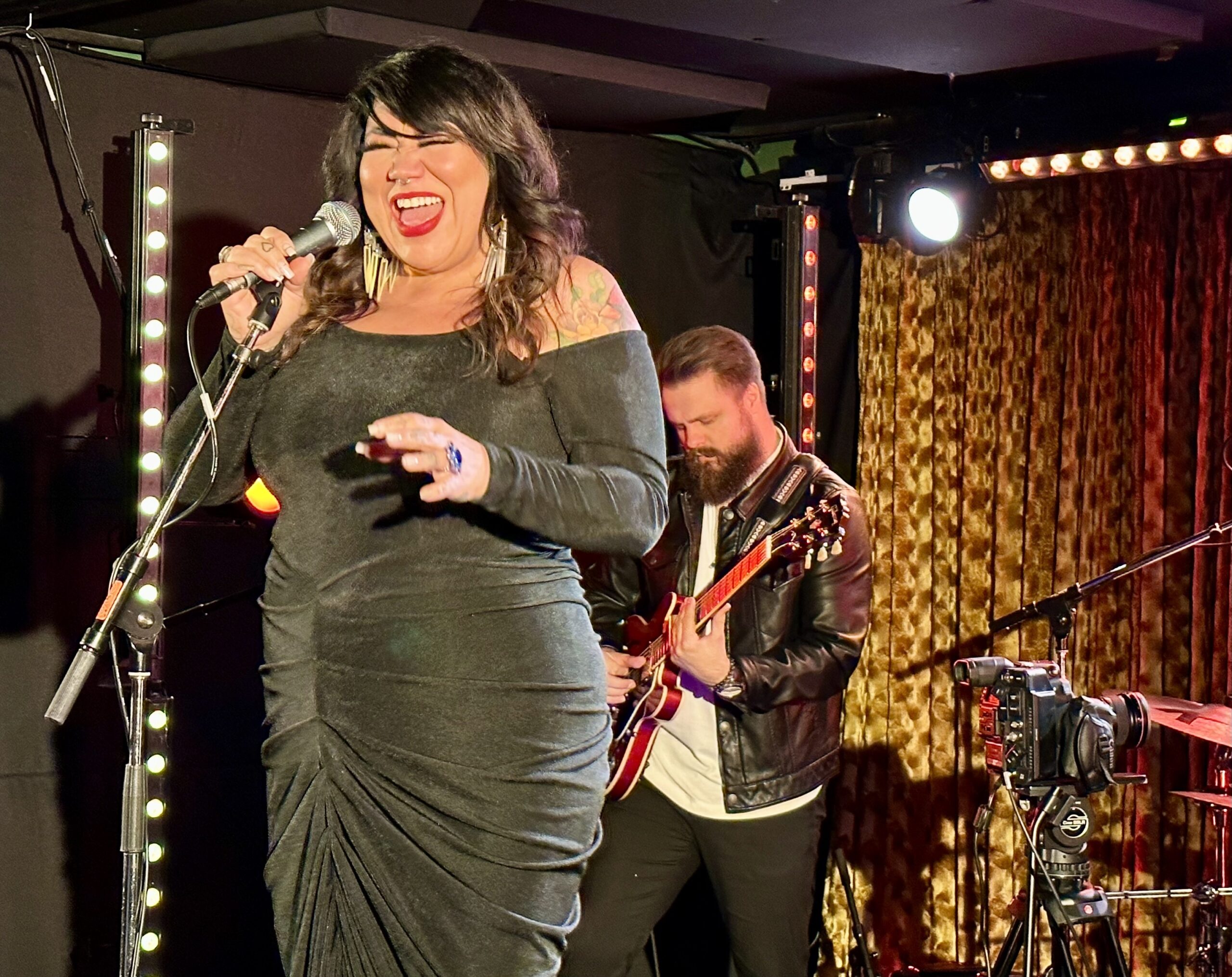 A woman in a black dress sings into a microphone as a bearded man plays guitar behind her on stage in a basement venue.