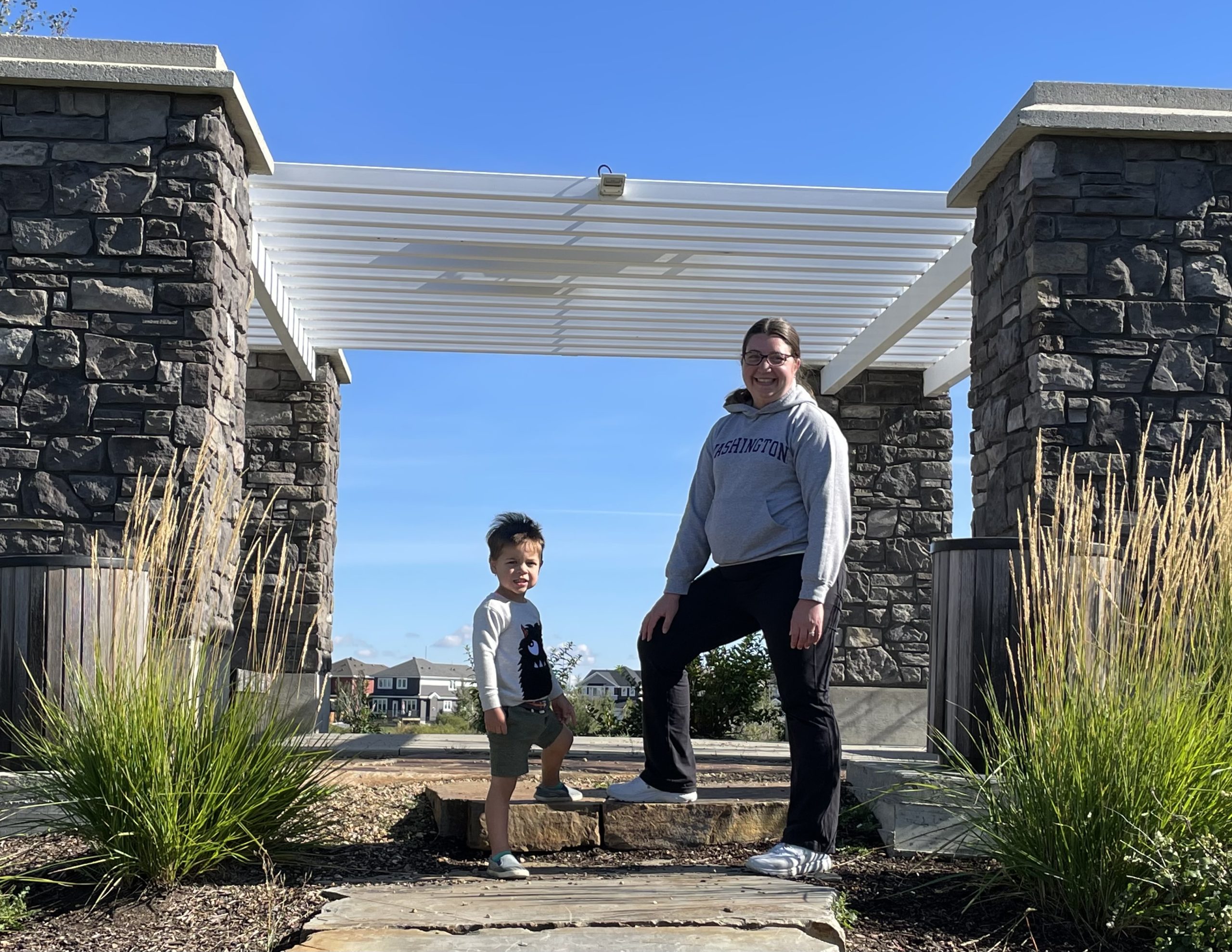 A woman and young boy stand in front of a pergola with stone pillars.