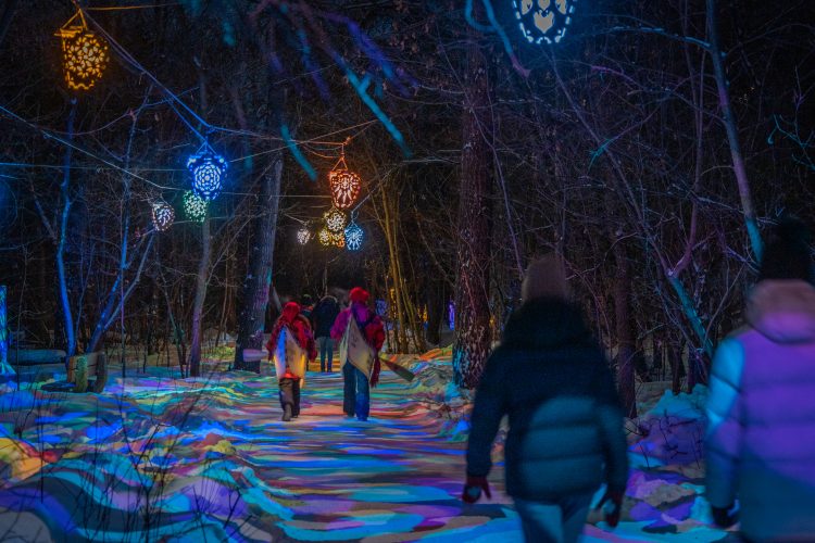 People walking along a pathway through trees in the winter at the Flying Canoë Volant Festival. The trees have colourful lights hanging.