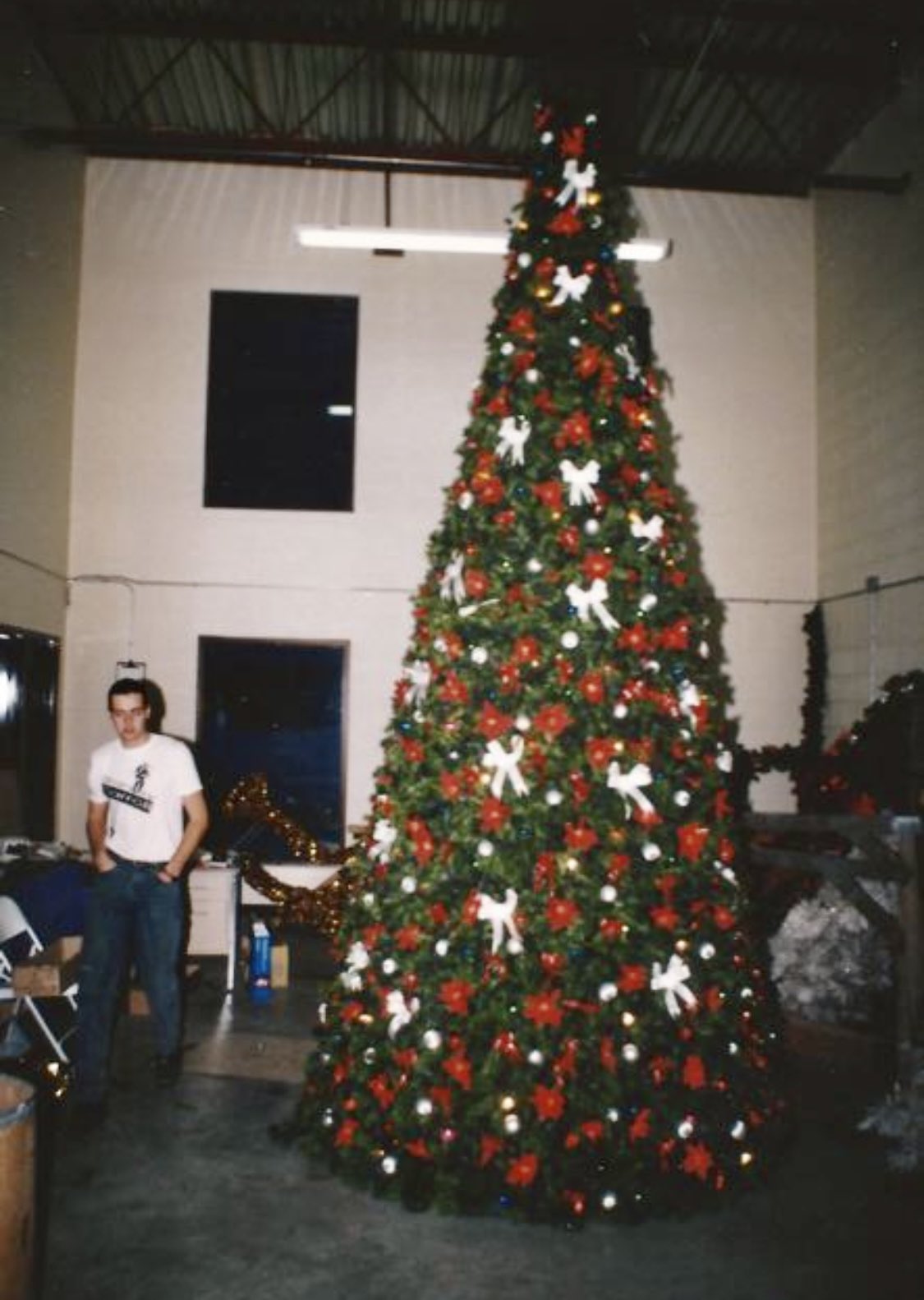 A man walks beside a tall Christmas tree decorated with red bulbs and white bows.