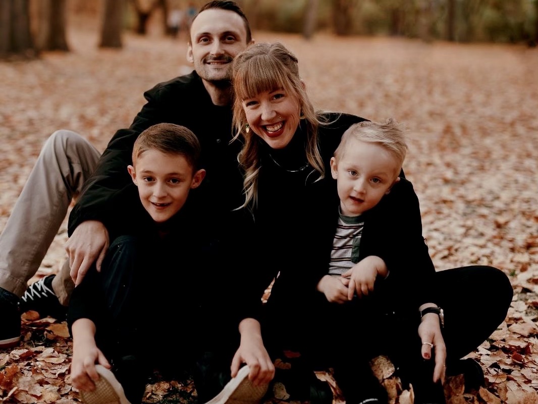 A man, woman and their two sons sit and smile in some leaves on a fall day in Edmonton.