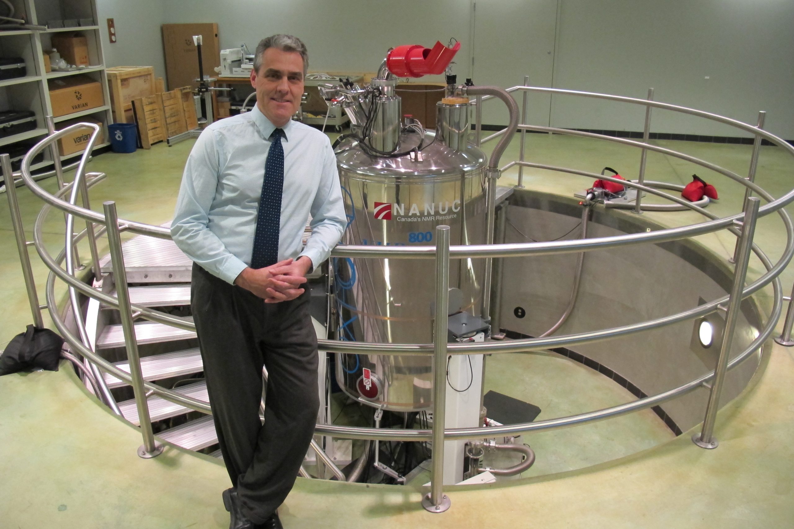 A man with his hands clasped in front of him leans on a railing that surrounds a spiral staircase and a large silver canister.