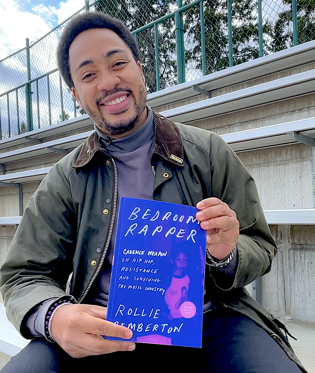 Rollie Pemberton sits in some bleachers holding a copy of his book, Bedroom Rapper.