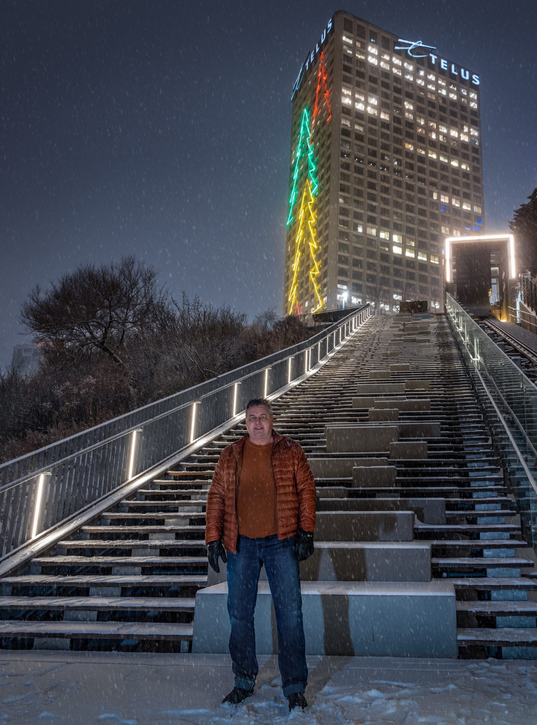 Slavo stands with the Telus building Christmas trees behind him at night during the winter. The trees are lit up in red, green and yellow.