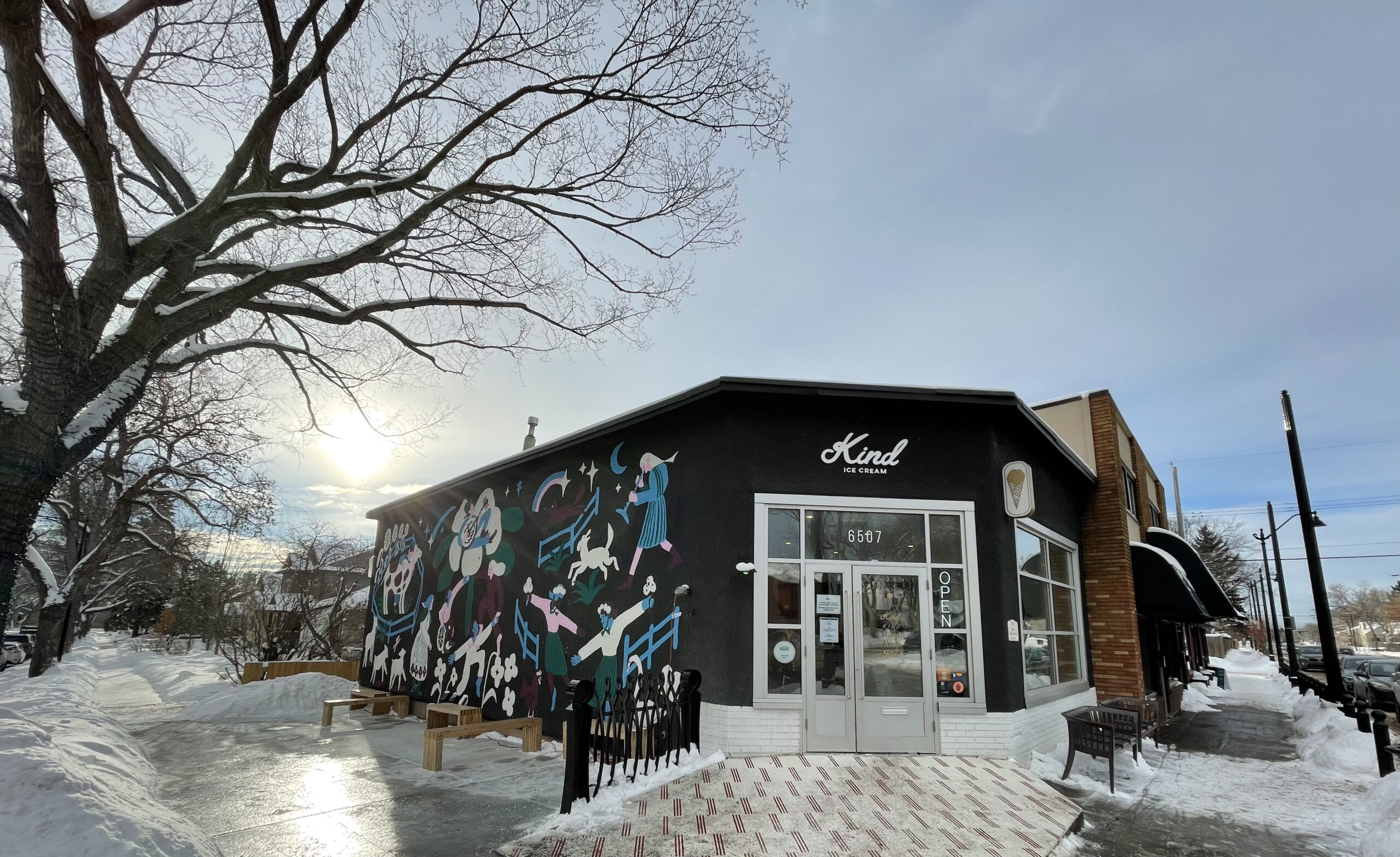 Kind Ice Cream Highlands location in 2022. There is snow on the ground and the sun shining. The front of the building says Kind Ice Cream and the side of the building has a mural.