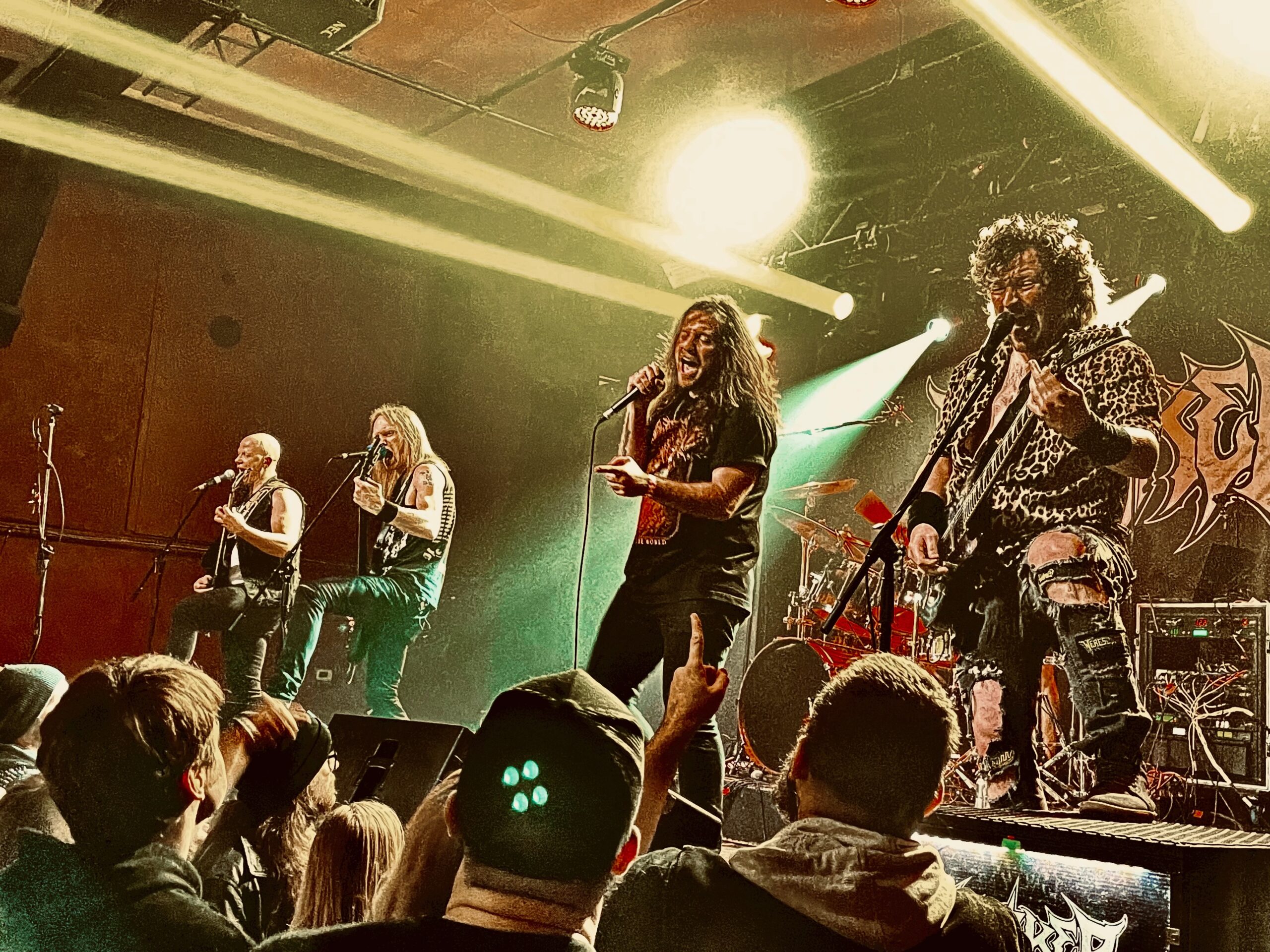 A group of five metal musicians perform on stage in front of some fans as yellow strobe lights stream over their heads.