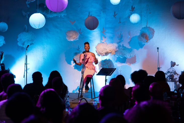 A woman sits on a chair as she performs her poetry on stage to an audience in a darkened room.