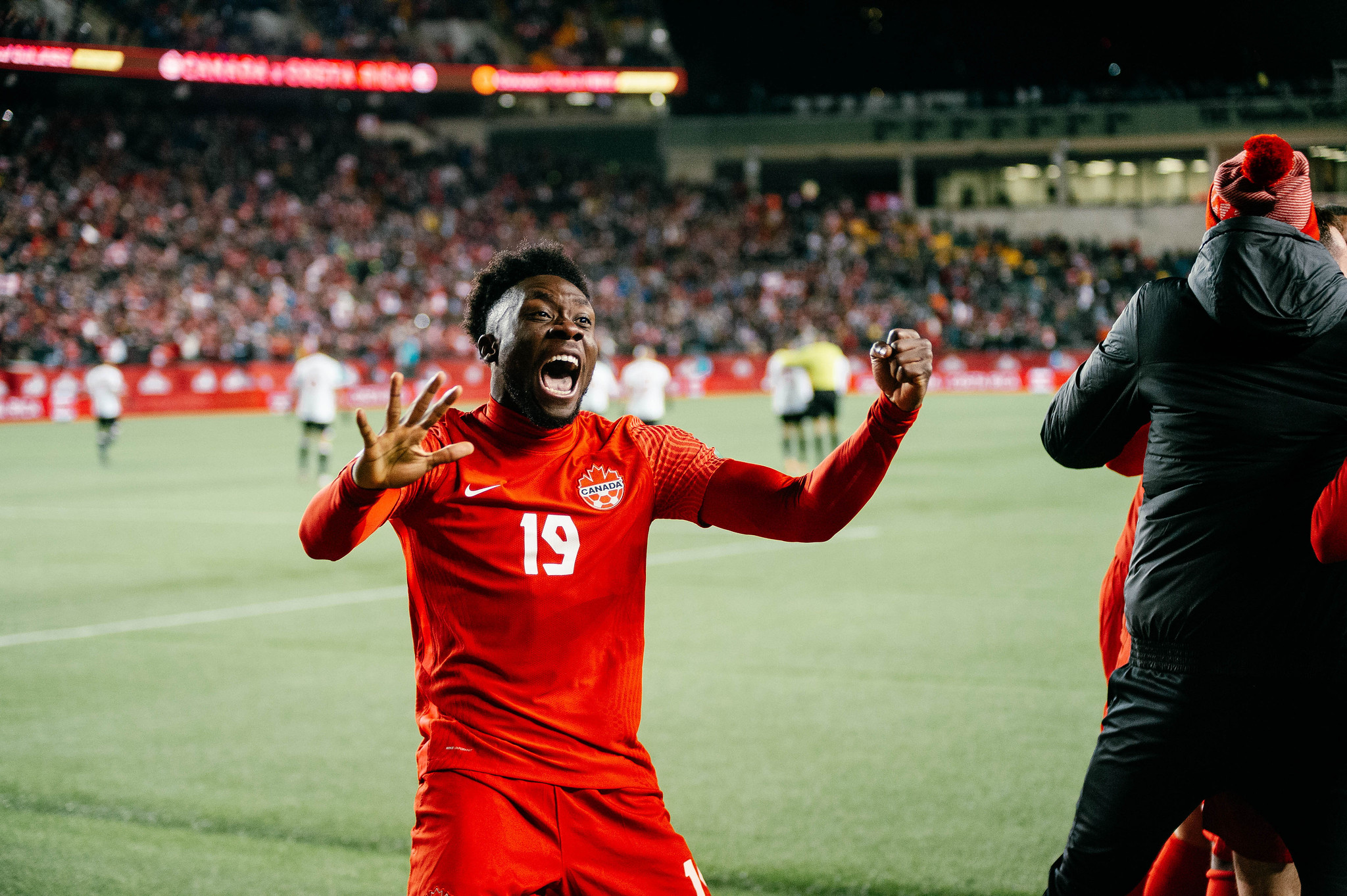 Alphonso Davies, wearing a red Canada soccer uniform, celebrates on the soccer field after a goal.