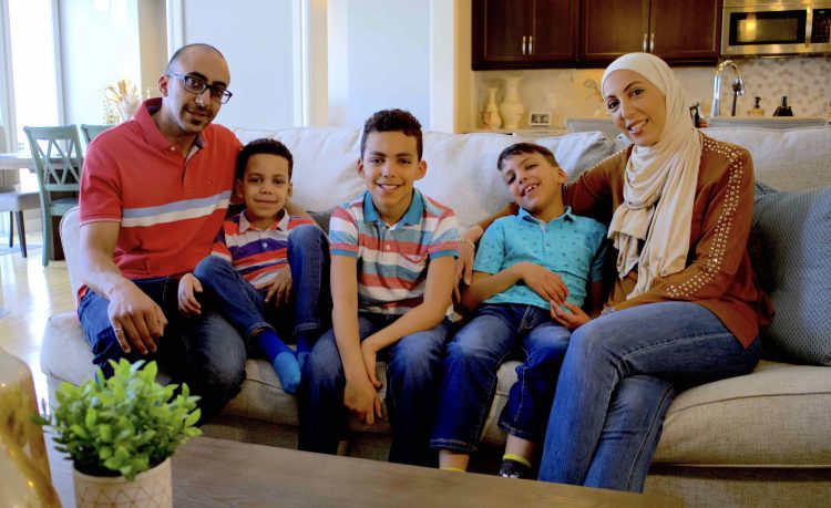 A family with 3 children sits on a couch inside their home.