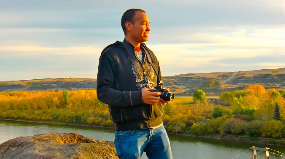 A man standing outdoors on a rocky outcrop, holding a camera and smiling. He is dressed in a black hoodie and blue jeans. The background features a scenic landscape with autumn trees and a river under a partly cloudy sky.