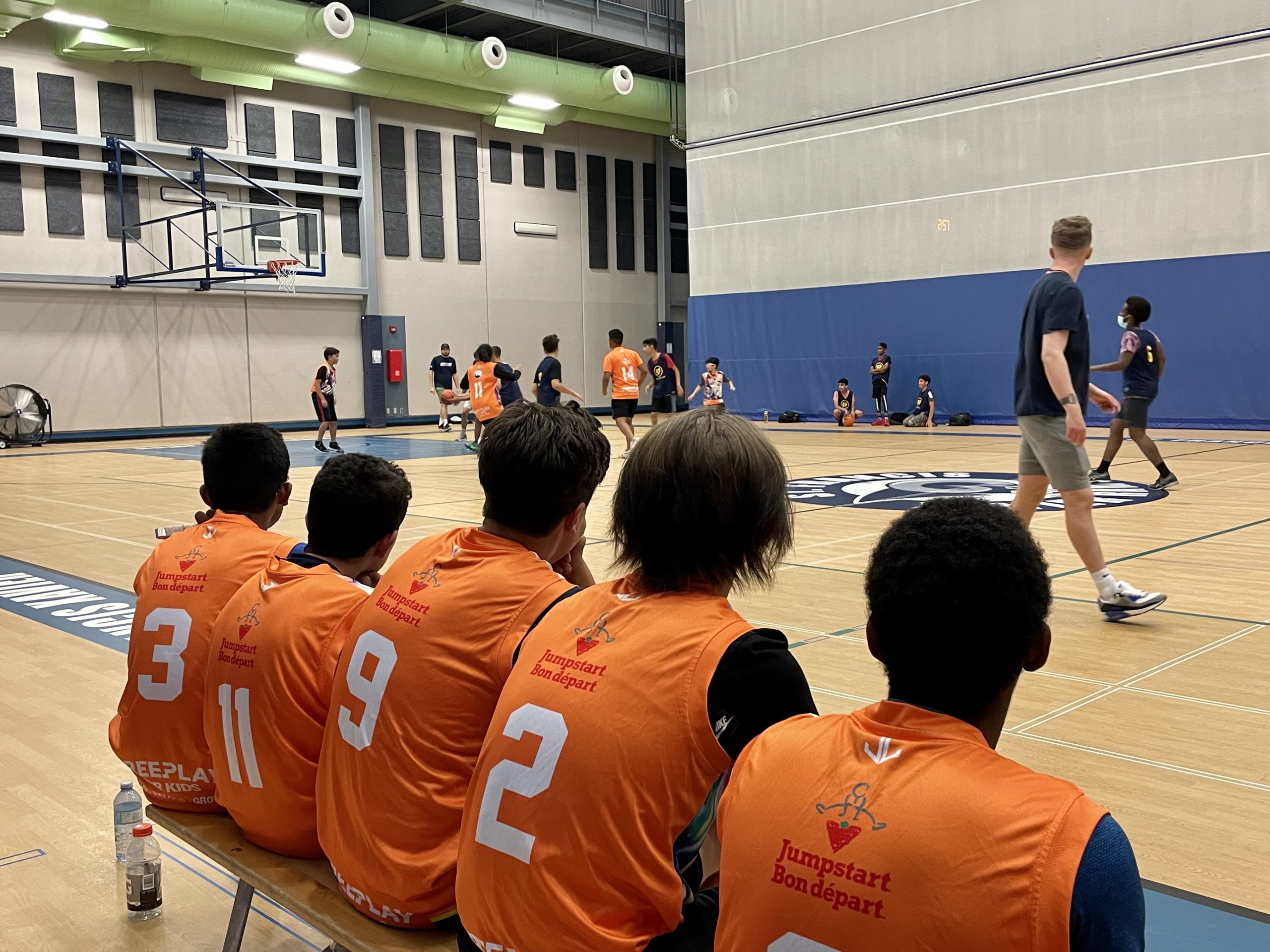 Five boys with their backs to the camera sit on a bench and watch a basketball game on the court in front of them.