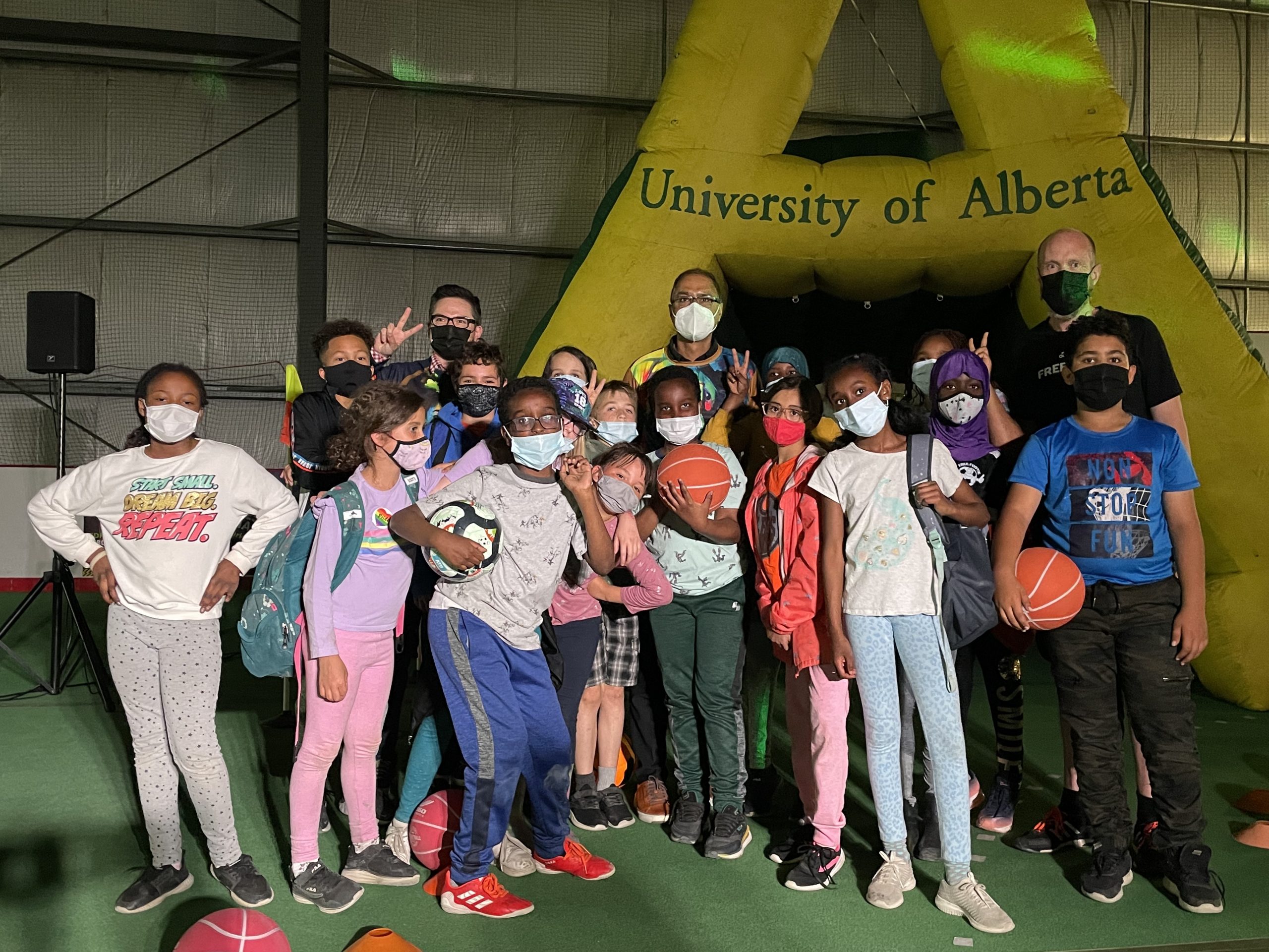 A group of children with masks and balls pose in front of three men on an indoor soccer field.