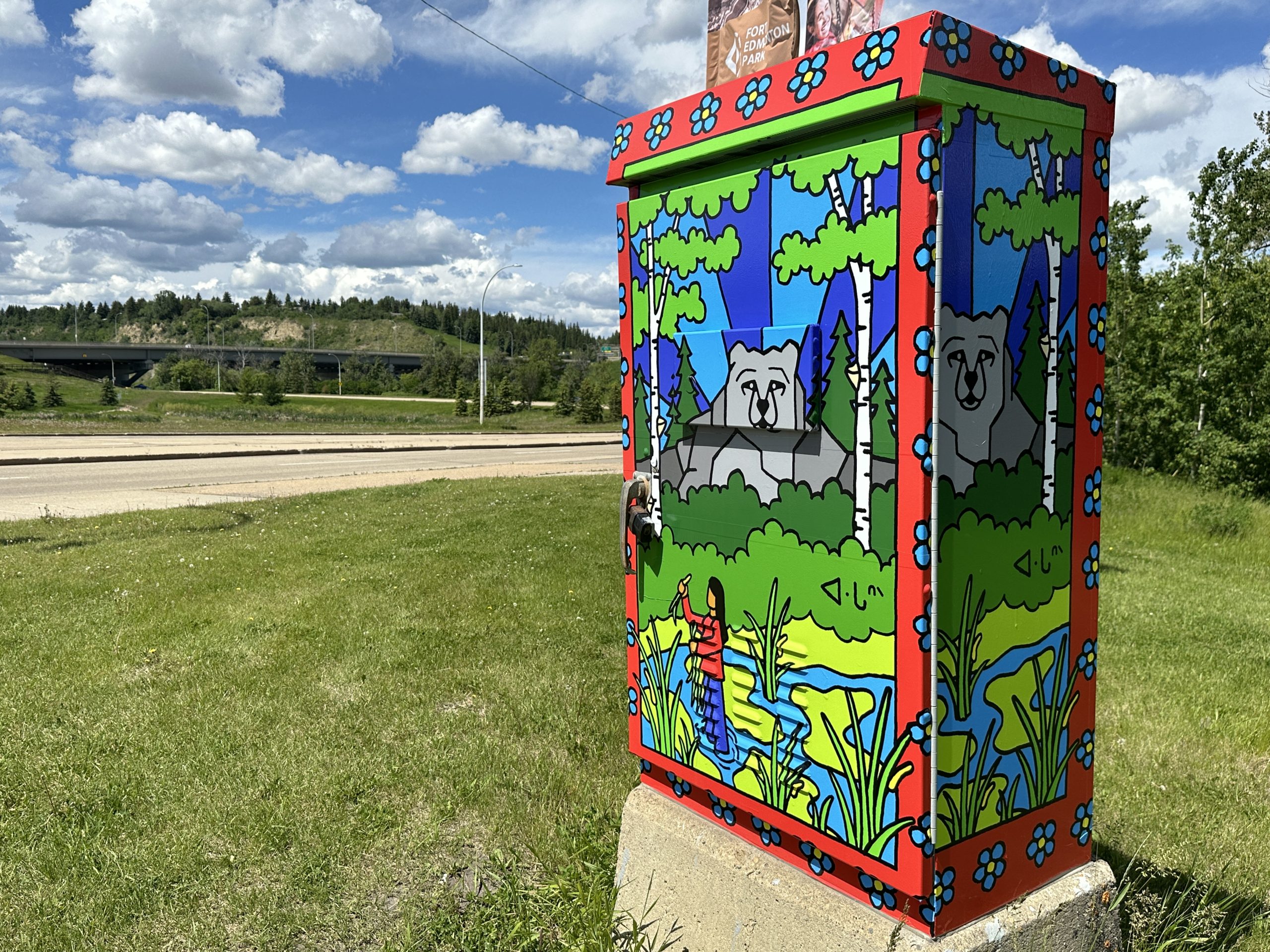 A control box wrapped with art of a bear in a forest and a person pulling rat root from a muskeg.