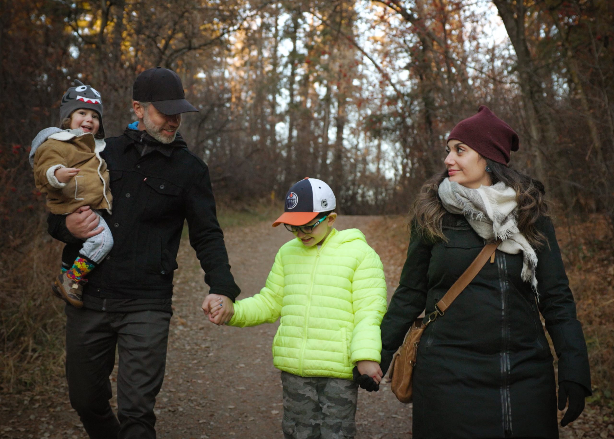 A father carrying a child walks holding hands with another child and mother on a forest pathway in fall.