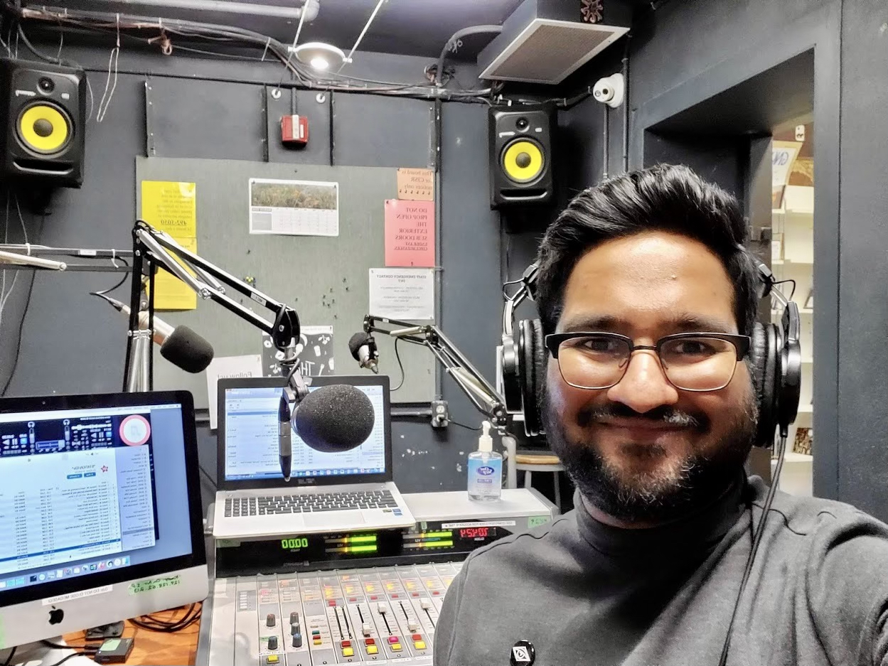 A smiling bearded man with glasses and headphones sits in front of a recording console, a computer screen, a laptop and a microphone.