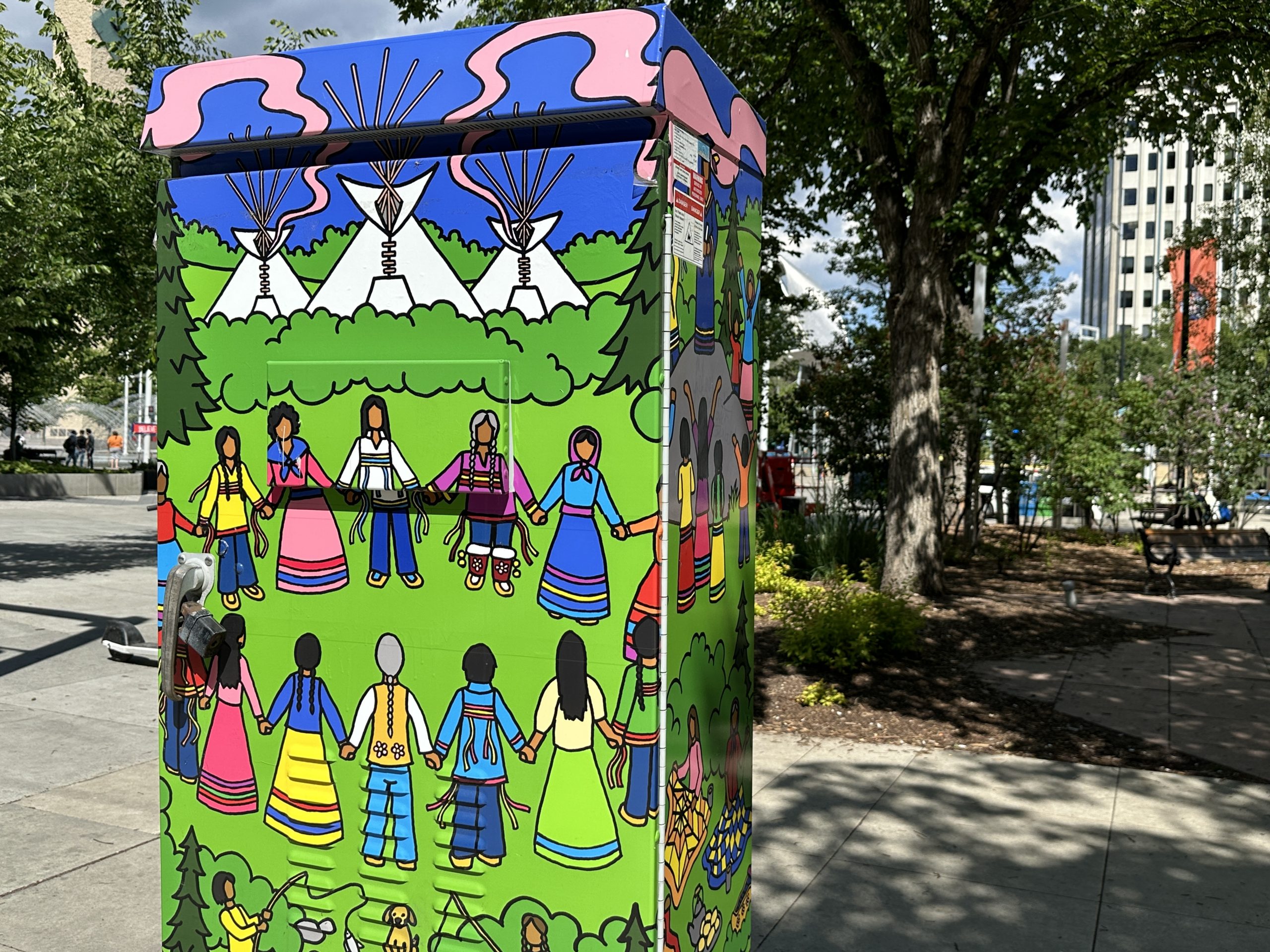 A traffic control box wrapped with art of a circle of Indigenous people holding hands in front of three tipis.