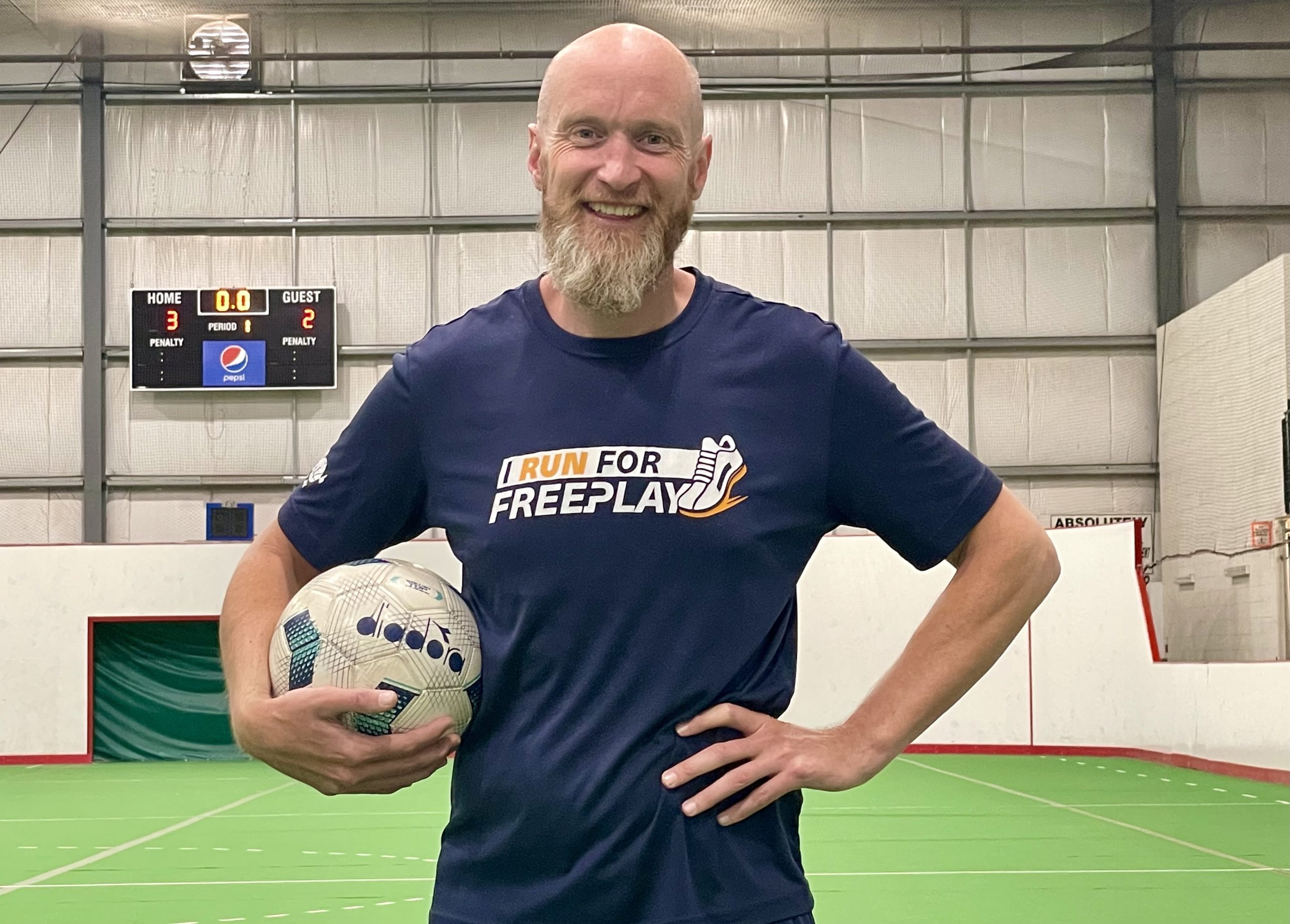 A bald man with a beard holds a soccer ball in his right hand as he stands on an indoor soccer field.